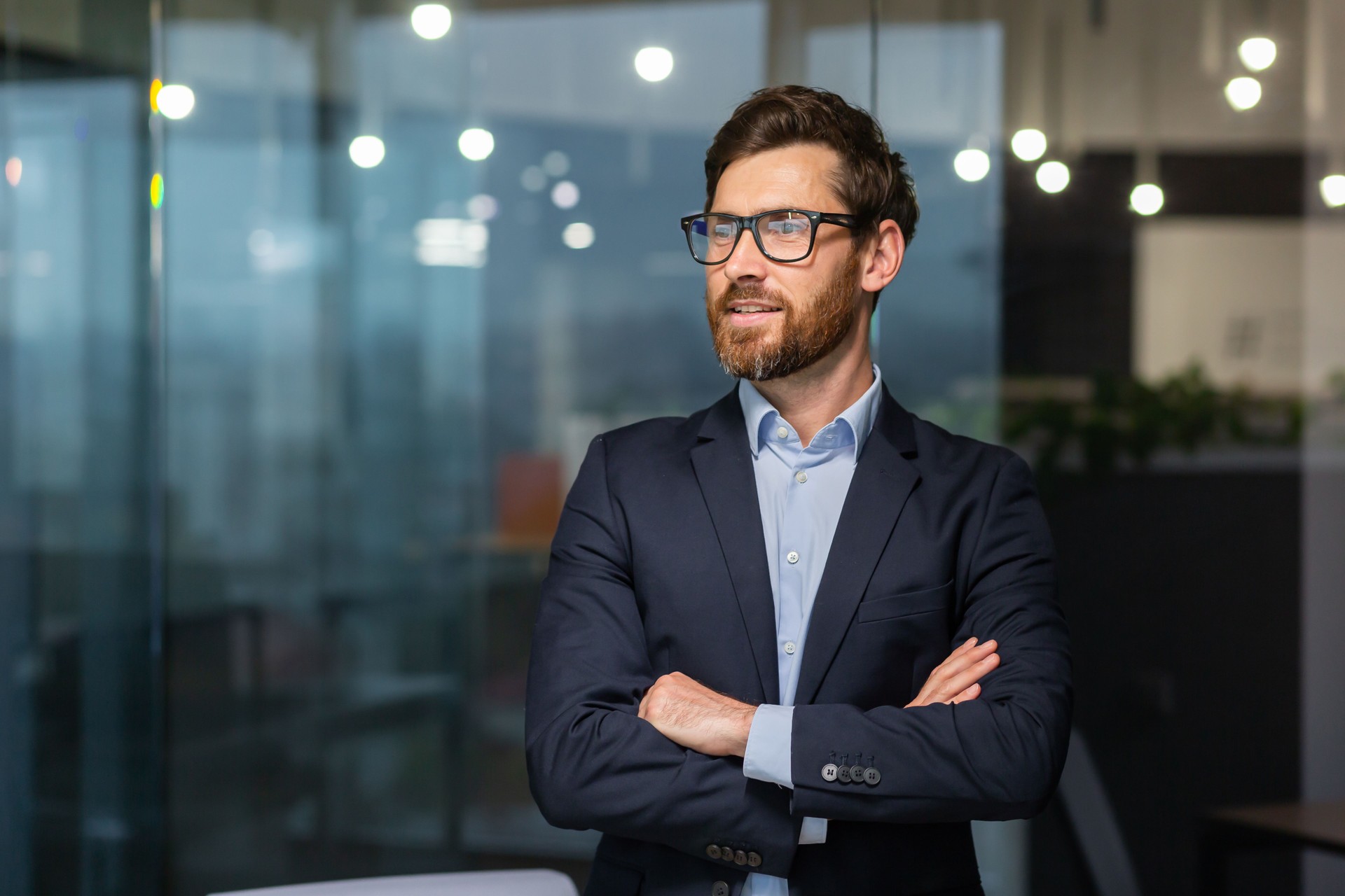Portrait of successful mature investor, senior businessman inside office looking towards window smiling with crossed arms, man in glasses and business suit